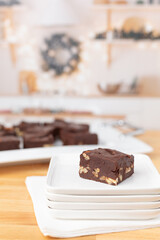 Piece of Homemade Chocolate Pecan Fudge on a White Plate with Tray of Fudge in Background in a Kitchen Decorated for Christmas