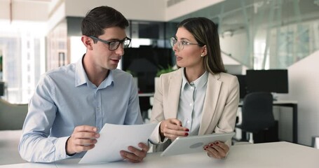 Two young European workmates discussing on-line presentation, comparing data, using digital tablet, holding papers, checking information in legal documents, engaged in teamwork in modern workspace