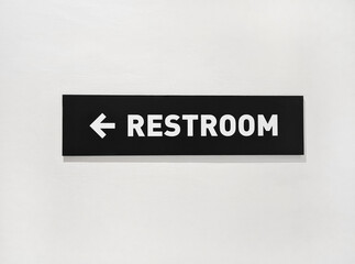 Restroom sign on a white wall. Path to Restroom with arrow. Black sign label indicating direction to Restroom in a room inside the building.