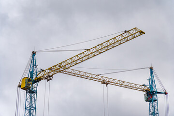 Two construction cranes are positioned at a construction site, actively working to lift heavy materials against a backdrop of overcast skies, indicating ongoing development efforts