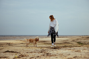 Young woman walking with dog on a sandy beach. Dog plays with a red ball, while the woman enjoys the calm and peaceful seaside atmosphere. Casual autumn clothing, plaid shirt tied around her waist.