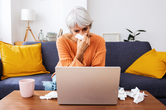 Senior sick woman sitting on sofa and blowing her nose with a tissue while having a video call with doctor from home