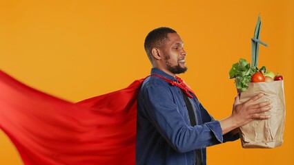 African american guy posing as a superhero with fresh ripe produce, hero with red cape promoting ethically sourced food and zero waste. Male model supports local farming. Camera B. © DC Studio