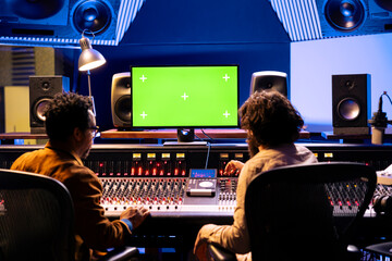 Team of artists mixing and mastering music with greenscreen on pc in professional studio, using audio console with switchers and sliders in control room. Singer and sound technician create songs.