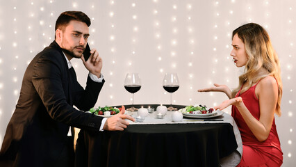 Relationship Problems. Angry Young Woman Blaming Her Busy Boyfriend Talking On Phone During Romantic Dinner In Restaurant, Side View