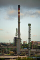 Fototapeta premium A telephoto shot of an industrial landscape with two tall smokestacks and complex refinery structures, surrounded by urban buildings. Overcast sky adds a moody, atmospheric touch to the scene