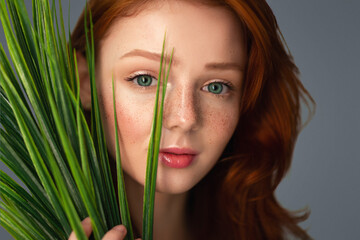Fototapeta premium Headshot Of Ginger-Haired Young Female Posing With Green Plant Looking At Camera Through Grass Over Gray Background. Studio Portrait. Beauty And Nature, Natural Facial Skin Care And Cosmetics