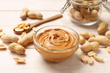 Tasty peanut butter in bowl, groundnuts, jar and spoon on wooden table, closeup