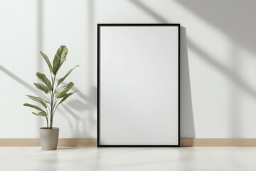 A black framed white picture sits on a wooden floor next to a potted plant