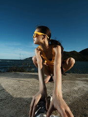 Woman, yellow shirt, sunglasses, sitting, water, outdoors a woman in a yellow shirt and sunglasses sits on the ground with her hands on her knees and feet on the ground, next to a body of water,