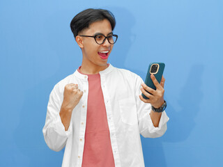 Happy young Asian man celebrates success while looking at his smartphone, raising hands in excitement on a blue background.