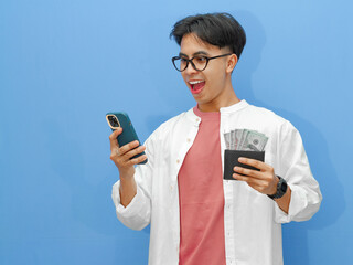 Portrait of a happy Asian man holding a wallet full of money and using a mobile phone with an...