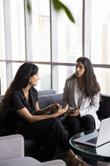 Serious young diverse businesswomen meeting in co-working for offline communication, talking on job interview, sitting on couch, discussing work project, teamwork strategy