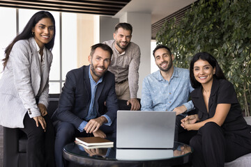 Diverse team of happy business colleagues posing for portrait in office co-working space. Multiethnic group of professionals meeting at laptop, sitting on couch at computer, looking at camera, smiling