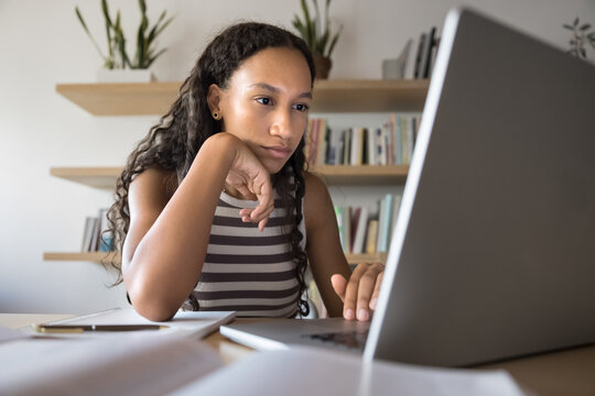Focused thoughtful young African student girl reading online article on laptop in public library, thinking on report for class, essay, touching chin, browsing Internet for research paper
