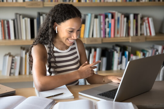 Cheerful young fresh Black college student girl talking on video conference call on laptop with earphones, smiling, laughing, using computer in library for online learning communication