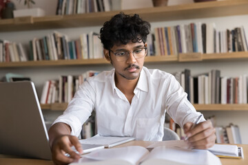 Focused smart male Indian student reading textbook in library, writing notes for essay, scientific article, research paper, sitting at workplace table with laptop, studying open book