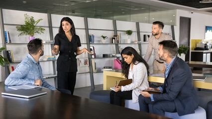 Confident young Middle Eastern business coach woman giving training to diverse employees writing notes. Female leader holding meeting with team in co-working workspace. Banner shot