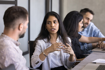 Serious beautiful young Indian professional woman talking to colleague at workplace. Multiethnic business colleagues, project managers talking at work table, discussing project tasks, teamwork