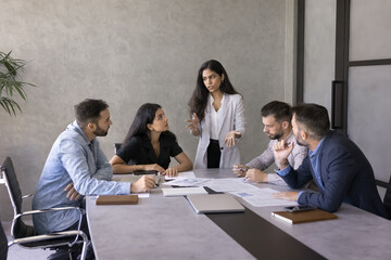 Young Indian business project manager woman presenting marketing reports, sales data to team, speaking to colleagues, standing at large meeting table. Diverse group analyzing financial research