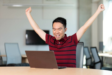 A college student sits at a table with arms raised in front of a laptop