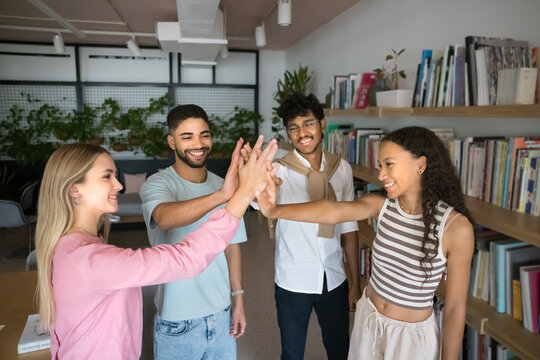 Diverse group of young students, college friends, classmates giving high five in university library space, standing at bookshelves, clapping hands in gesture of teamwork success, achievement