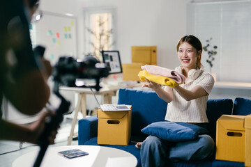 Online seller is recording a video of herself presenting clothes while sitting on her couch, with boxes and a laptop in the background