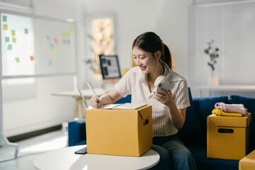 Young asian woman is smiling while writing on a notepad and looking at her phone in her home...