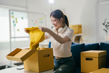 Young woman is unpacking a yellow sweater from a cardboard box. She is excited to be moving into...