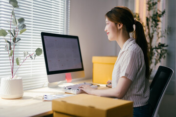 Asian entrepreneur happily works from her home office, checking orders for her online store on her computer in a bright, inviting workspace