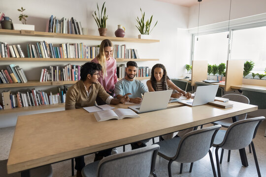 Multiethnic students doing college homework in library together, working on group project for class, discussing research study, using laptops, sitting at table with bookshelves in background - Powered by Adobe
