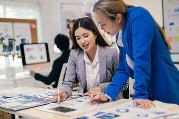 Fototapeta premium Two businesswomen in a modern office discuss finance and marketing, surrounded by charts and graphs. They collaborate, plan, and analyze data, showing teamwork and professionalism