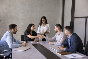 Happy successful female business leader shaking hands with employee at corporate meeting table. Diverse coworkers, partners ending negotiation with handshake, smiling, laughing