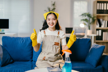 Cheerful asian woman wearing an apron and yellow gloves is showing a thumbs-up gesture while sitting on a sofa in the living room