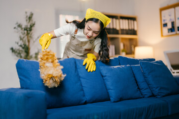 Young maid wearing yellow gloves is cleaning a blue sofa with a feather duster, she is working hard...