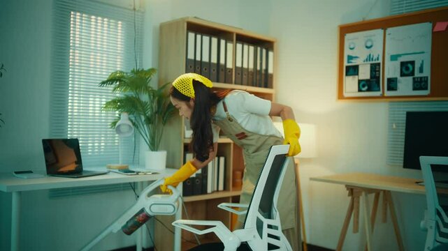 Young woman wearing an apron and gloves is vacuuming an office chair. She is working diligently to ensure the office is clean and presentable