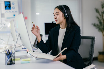 Asian businesswoman is working in an office setting, wearing a headset and using a computer while holding a pen and notepad. She is fully engaged with her work