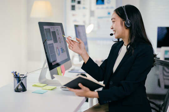 Asian businesswoman wearing headset is analyzing financial charts on computer and having a video conference call with client - Powered by Adobe