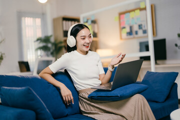 Obraz premium Young asian woman is working remotely from home, sitting on a blue sofa and having a video call on her laptop. She is wearing headphones and gesturing with her hand as she speaks