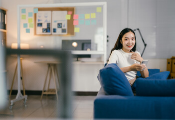 Fototapeta premium Young asian woman takes a break, happily browsing social media on her phone in a modern living room. The blue and white decor adds to her cozy work-from-home day