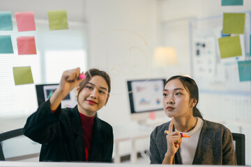 Two young businesswomen are brainstorming, using a glass board and sticky notes to organize their ideas in a modern office - Powered by Adobe