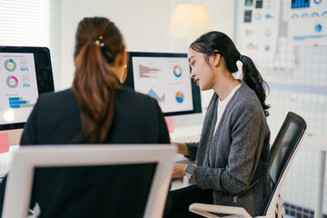 Two asian businesswomen are working together in the office. They are looking at charts and graphs on a computer screen, analyzing financial data