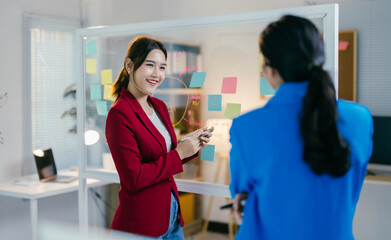 Two businesswomen are having a productive meeting in the office, using sticky notes on a glass wall to brainstorm ideas