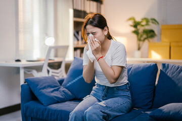 Young woman is blowing her nose with a tissue while sitting on a sofa in her living room, suggesting she may be feeling unwell