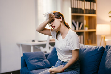 Unwell asian woman sits on her couch, wiping sweat and holding an ice pack to her head, showing...