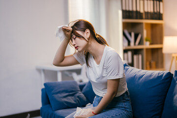 Young woman is wiping sweat from her forehead with a tissue while sitting on her sofa at home,...