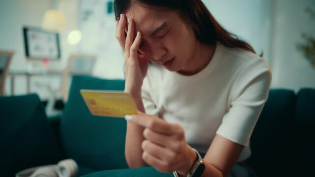 Young asian woman sitting on the sofa at home, feeling overwhelmed by financial stress while holding her credit card. Worries about expenses and payments create anxiety and frustration