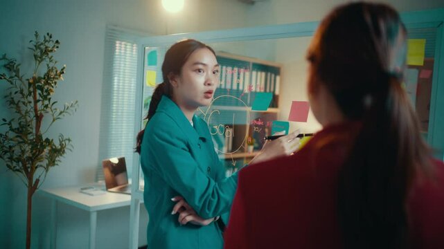 Businesswoman explains strategy to a colleague using a glassboard and sticky notes, fostering collaboration and discussion in a professional office environment