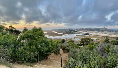 A winding dirt trail leads down to the San Elijo Lagoon at sunset, with dramatic clouds, coastal...