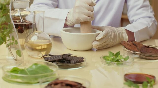 In a template video taken in a laboratory from the top viewpoint, a scientist who is hooked to herbs puts it in a white mortar. The scientist is surrounded by glasses filled with liquids and plants.